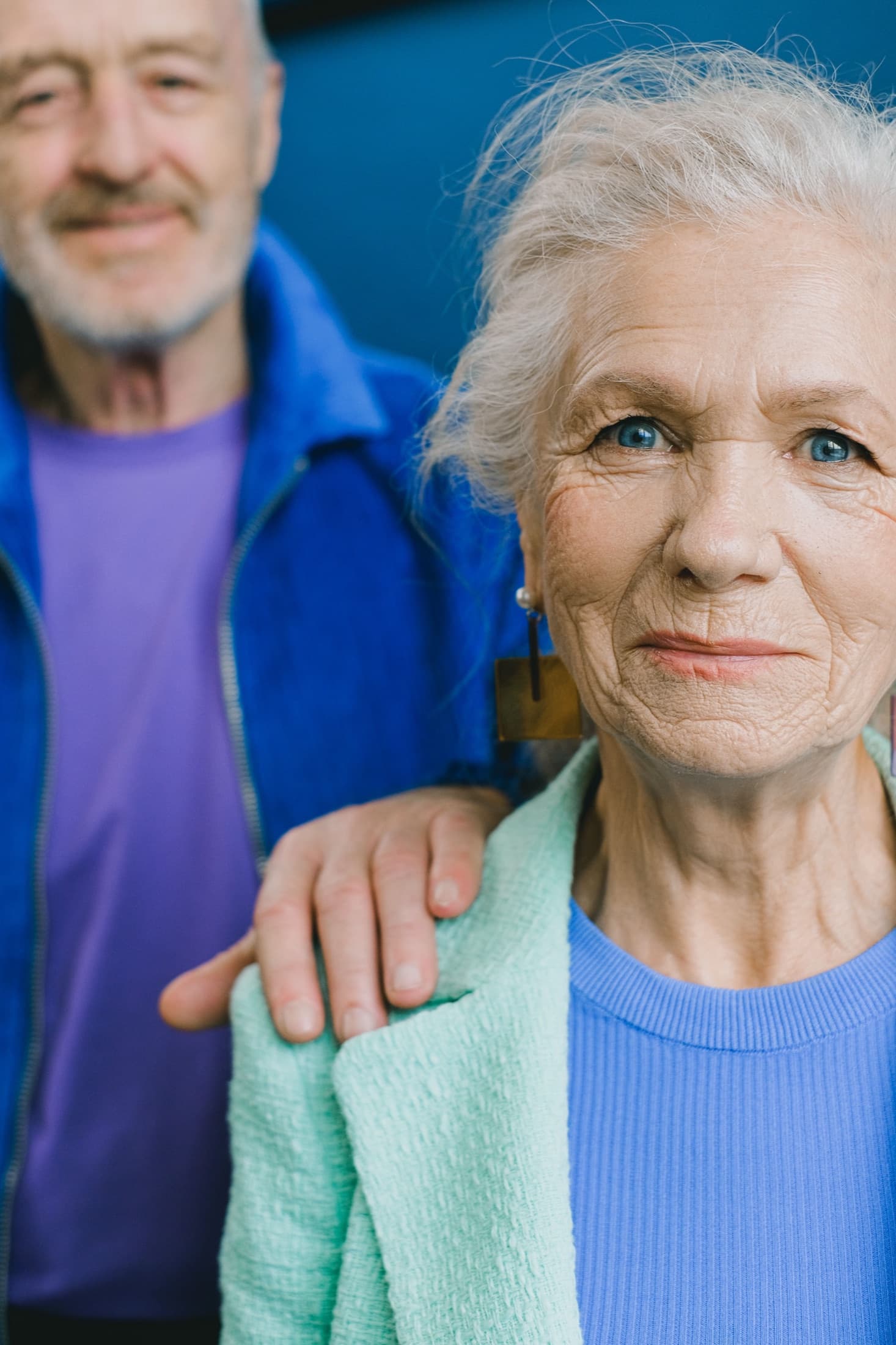 An elderly couple together, the husband's hand resting on his wife's shoulder
